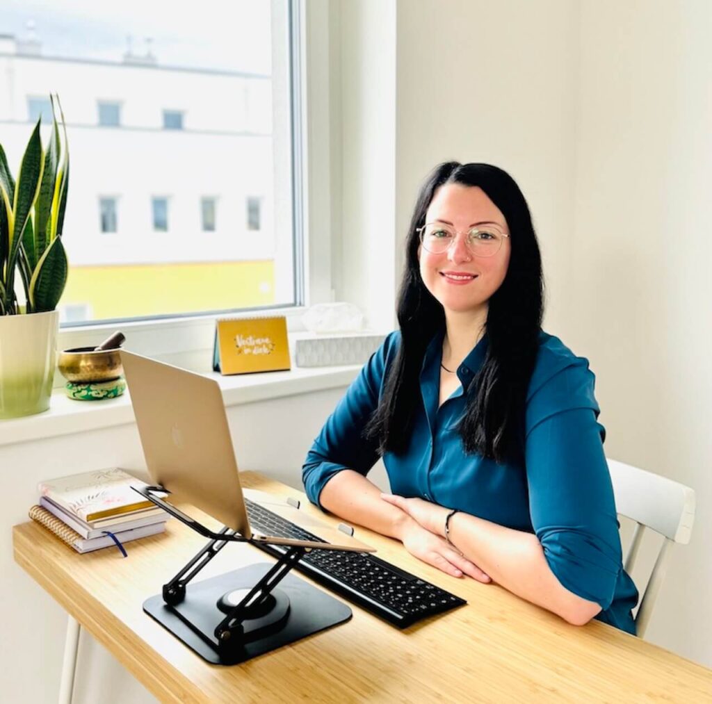Clinical online psychologist Angelika Spies in her office - friendly look at the camera, seated at desk with laptop - psychological therapy and counselling via video, audio and chat